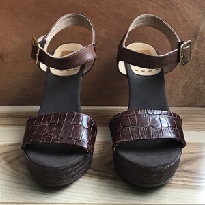 May include: A pair of brown leather platform sandals with an ankle strap and buckle closure. The sandals have a crocodile-embossed design.