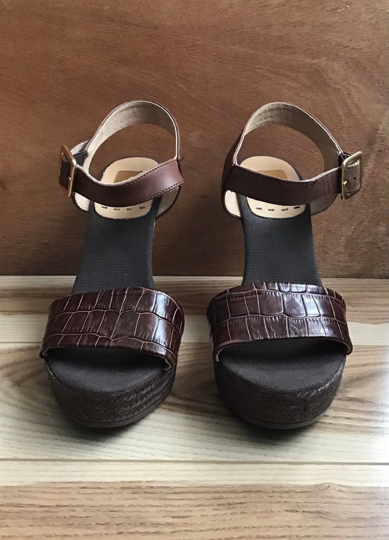 May include: A pair of brown leather platform sandals with an ankle strap and buckle closure. The sandals have a crocodile-embossed design.