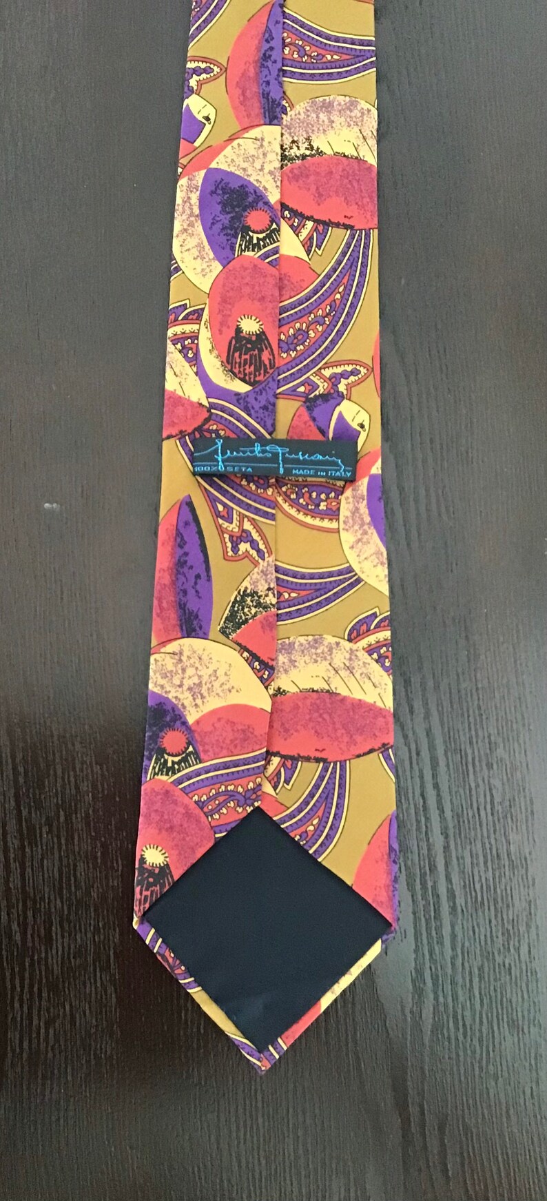 May include: A brown silk tie with a paisley pattern in shades of purple, pink, and yellow. The tie has a black label that reads "Frank  Frasconi 100% Seta Made in Italy".