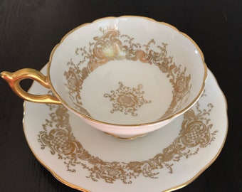 May include: A white teacup and saucer with a gold rim and intricate gold floral pattern.