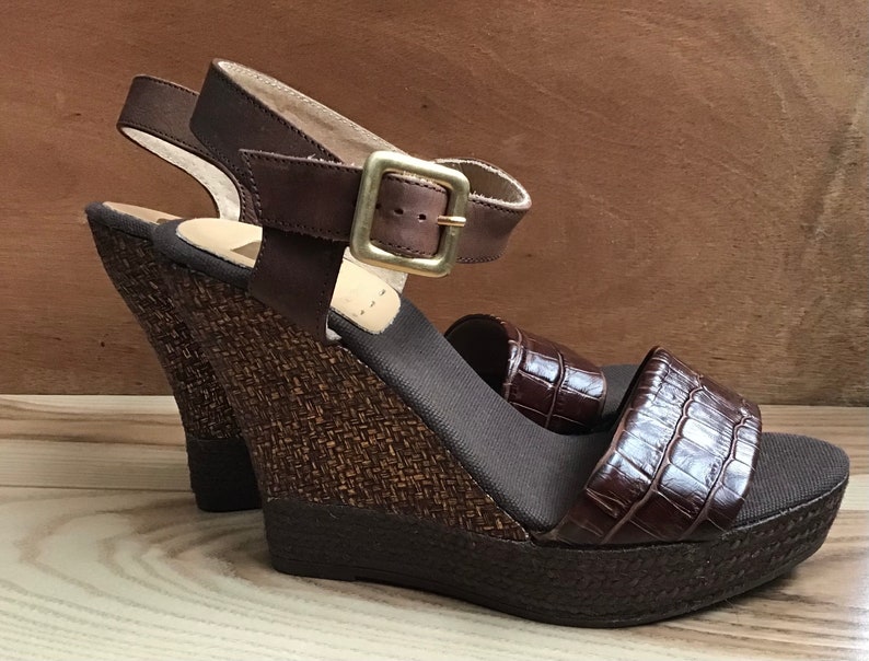 May include: Brown leather wedge sandals with a woven platform and a buckle closure. The sandals have a crocodile-embossed leather strap.