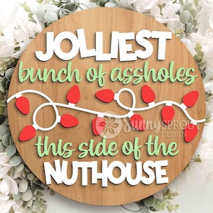 May include: A round wooden sign featuring the white text JOLLIEST, the green text bunch of assholes, a string of red Christmas lights, the green text this side of the, and the white text NUTHOUSE. The sign includes a Sunny Sprout Design logo.