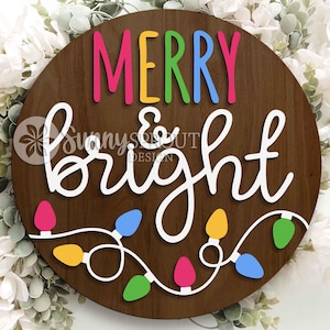 May include: A circular dark wood sign featuring the text MERRY in multi-coloured letters and bright in white cursive script. Below the text is a string of colourful light bulbs in pink, blue, yellow, and green. The sign includes a watermark logo.