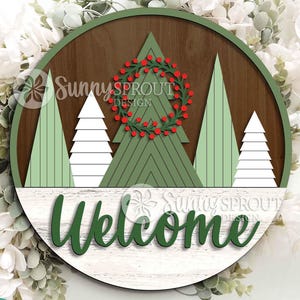 May include: A circular wooden wall sign featuring a dark brown background with layered green and white pine trees. A red berry wreath is centered on the largest tree. The word Welcome is written in dark green script over a white wood grain base.
