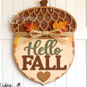 May include: A wooden acorn-shaped door sign featuring a dark brown cap with a scalloped pattern, a cream-coloured base, and the text "Hello Fall" in green and brown. It is decorated with two orange fabric leaves and a jute twine bow.