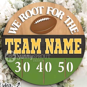 May include: A circular wooden sign featuring a brown football graphic and the white text "WE ROOT FOR THE" above the yellow text "TEAM NAME" on a dark background, with a green field graphic at the bottom displaying the white numbers 30, 40, and 50.