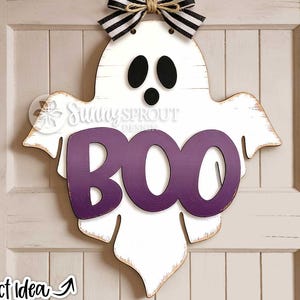 May include: A white wooden ghost-shaped wall hanging featuring black eyes and mouth, with the purple text BOO centered on the front. A black and white striped fabric bow is attached to the top, and the edges show a distressed brown finish.