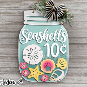 Puede incluir: Un letrero de madera con forma de tarro con la frase "Seashells 10¢". El tarro contiene recortes de conchas marinas de colores, una estrella de mar y un lazo. El letrero está decorado con vegetación.