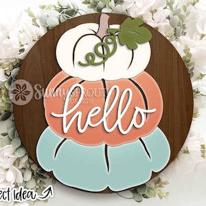Puede incluir: Un letrero de madera con tres calabazas apiladas en blanco, naranja y verde azulado. La palabra "hello" está escrita en escritura blanca sobre la calabaza del medio. Una hoja y una vid verdes acentúan la calabaza superior.