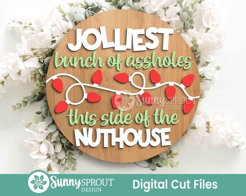 May include: A round wooden sign featuring the white text JOLLIEST, the green text bunch of assholes, a string of red Christmas lights, the green text this side of the, and the white text NUTHOUSE. The sign includes a Sunny Sprout Design logo.