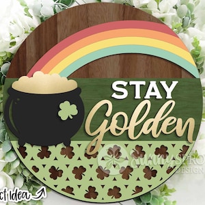 Op de afbeelding: Een rond houten bord met een groene achtergrond en een gouden pot met goud bovenaan. Een regenboog boogt over de pot. De woorden "STAY GOLDEN" staan in gouden letters op het bord. De onderkant van het bord is versierd met een patroon van groene klavertjes.