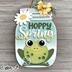 May include: A wooden jar-shaped sign with a frog design and the words "Hoppy Spring." The sign features a daisy, a yellow bow, and greenery at the top. The frog is green with black eyes and a smiling face. The sign hangs from a twine string.