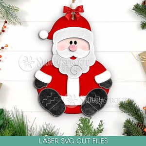 May include: A wooden Santa Claus ornament with a red suit, white beard, and black boots. The ornament has a red hat with a white trim and a small red bow at the top. The text "LASER SVG CUT FILES" is at the bottom.