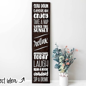 May include: A dark brown wooden sign with white text that reads "Slow Down Daydream Enjoy Take a Nap Watch the Sunset Relax Appreciate Today Laugh Read a Book Unwind Sip a Drink".