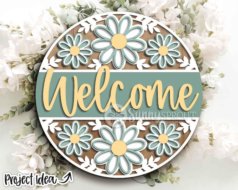 May include: A round wooden sign with a daisy theme. The sign has a brown background, a teal banner with the word "Welcome" in yellow script, and white daisy cutouts. The sign is surrounded by white flowers.