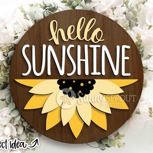 May include: A circular wooden sign featuring the words "hello SUNSHINE" in white and yellow. A yellow and black sunflower design is at the base. The sign is set against a backdrop of white flowers and foliage.