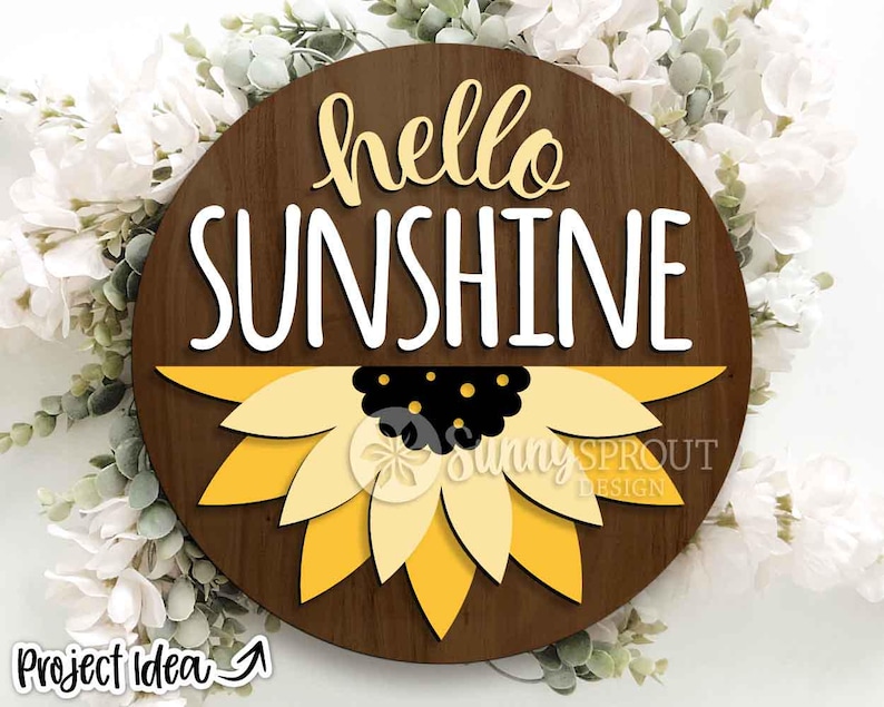 May include: A circular wooden sign featuring the words "hello SUNSHINE" in white and yellow. A yellow and black sunflower design is at the base. The sign is set against a backdrop of white flowers and foliage.