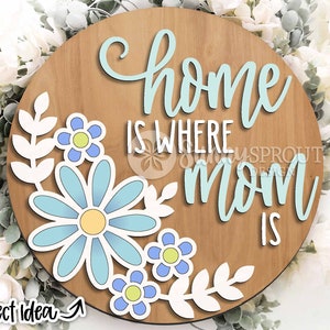 May include: A round wooden sign with the text "Home is where Mom is" in light blue lettering. The sign is decorated with white paper leaves and blue flowers.