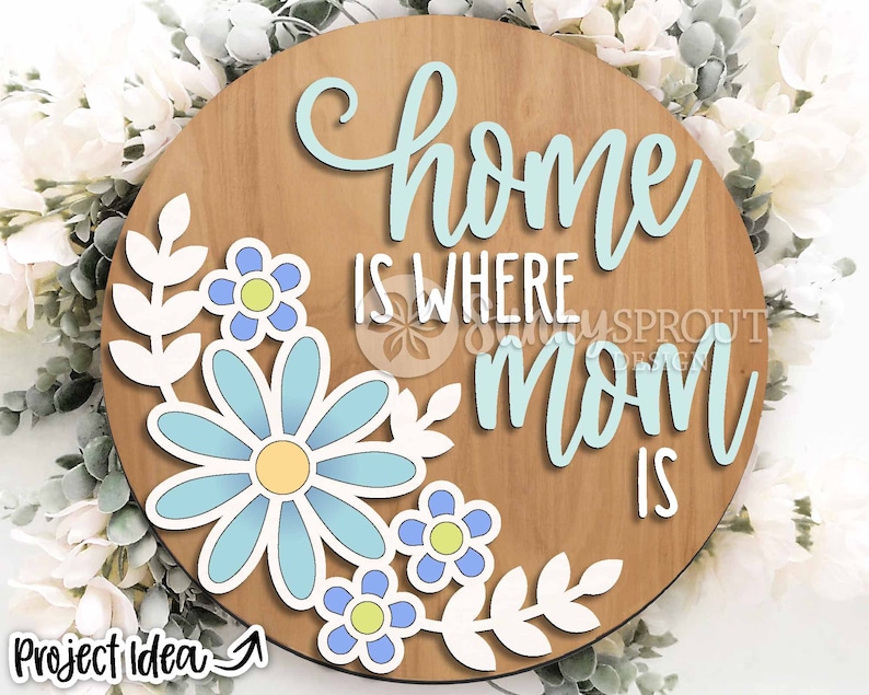 May include: A round wooden sign with the text "Home is where Mom is" in light blue lettering. The sign is decorated with white paper leaves and blue flowers.