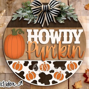 May include: A round wooden door sign featuring the text HOWDY PUMPKIN in white and orange lettering. The sign includes a large orange pumpkin graphic, a black and white cow print pattern with small orange pumpkins, and a black and white striped bow.