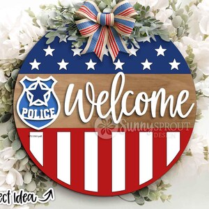 May include: A circular wooden sign with a patriotic design. It features a blue section with white stars, a wood-effect middle, and red and white stripes at the bottom. The word "Welcome" is in white script. A police badge is on the left. A red, white, and blue bow is at the top.