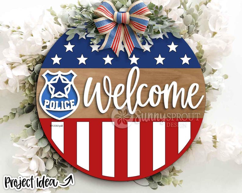 May include: A circular wooden sign with a patriotic design. It features a blue section with white stars, a wood-effect middle, and red and white stripes at the bottom. The word "Welcome" is in white script. A police badge is on the left. A red, white, and blue bow is at the top.