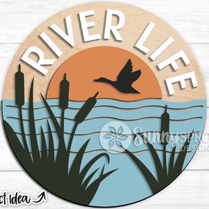 May include: A round wooden sign with the text "River Life" in white letters. The sign features a silhouette of a duck flying over a blue lake with cattails and a sunset in the background.