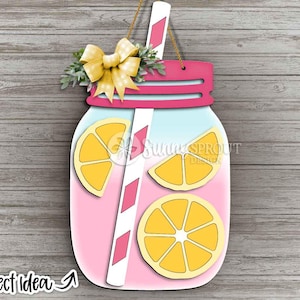 May include: A decorative wooden wall hanging shaped like a mason jar filled with pink lemonade, featuring three yellow lemon slices, a white straw with pink diamond patterns, a yellow gingham bow, and a pink lid, suspended by a thin brown rope.