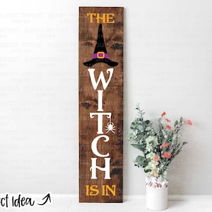 Peut inclure: Une pancarte en bois avec le texte "The Witch Is In" en lettres blanches. Un chapeau de sorcière noir avec un bandeau violet est au-dessus du texte. La pancarte est brune avec une texture de grain de bois.