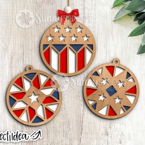 May include: Three wooden Christmas ornaments with patriotic designs. One ornament features stars and stripes, while the others have star and geometric patterns in red, white, and blue. A red bow and string are attached to the top ornament.