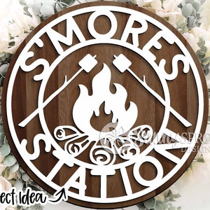 May include: White wooden sign with the words "S'mores Station" in a circle. The centre of the sign features a white silhouette of a campfire with flames and marshmallows on sticks.
