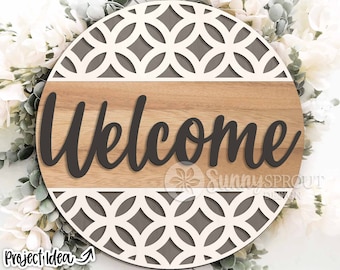 May include: Round wooden welcome sign with a gray and white geometric pattern. The word "Welcome" is cut out in black lettering.