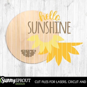 May include: A circular wooden sign featuring the words "hello SUNSHINE" in yellow and brown. A yellow and brown sunflower design is on the right. The sign is set against a white wooden backdrop.