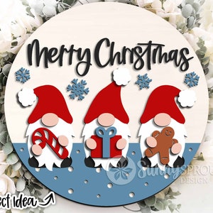 May include: A round wooden sign with the text "Merry Christmas" in black script. Three gnomes are depicted, each holding a different Christmas-themed item: a candy cane, a gift box, and a gingerbread man. The gnomes are red, white, and brown, and the background is blue with white snowflakes.