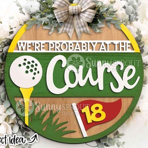 以下が含まれることがあります： 「WE'RE PROBABLY AT THE COURSE」の文字が入った丸い木製サイン。ティーに乗ったゴルフボール、18番の赤い旗、緑の芝生が特徴です。装飾的なリボンと花の配置が上部にあります。