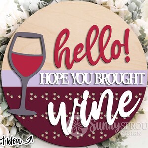 May include: Round wooden sign with the text "hello! hope you brought wine" in red and white lettering. A glass of red wine is depicted on the left side of the sign.