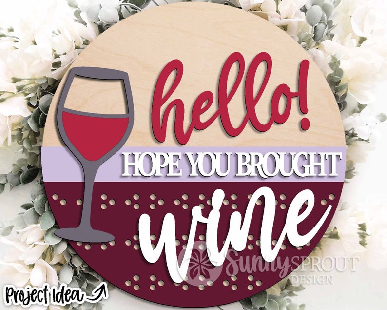 May include: Round wooden sign with the text "hello! hope you brought wine" in red and white lettering. A glass of red wine is depicted on the left side of the sign.