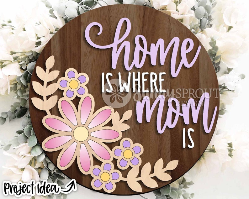 May include: A round wooden sign with the text "home is where mom is" in purple lettering. The sign is decorated with a floral design featuring pink and purple flowers with yellow centers.