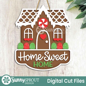 May include: A brown gingerbread house-shaped sign with white accents, a red heart, and a candy swirl. The sign reads "Home Sweet Home" in white script with green lettering. The sign is hanging by a gold string.