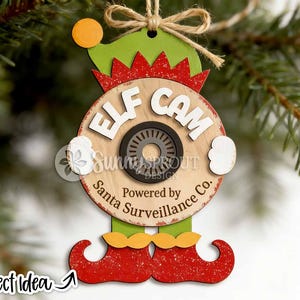 May include: A wooden Christmas ornament shaped like an elf with a green hat, red glittery shoes, and a central circular disc featuring the text ELF CAM and Powered by Santa Surveillance Co. with a black camera lens graphic and a jute twine hanger.