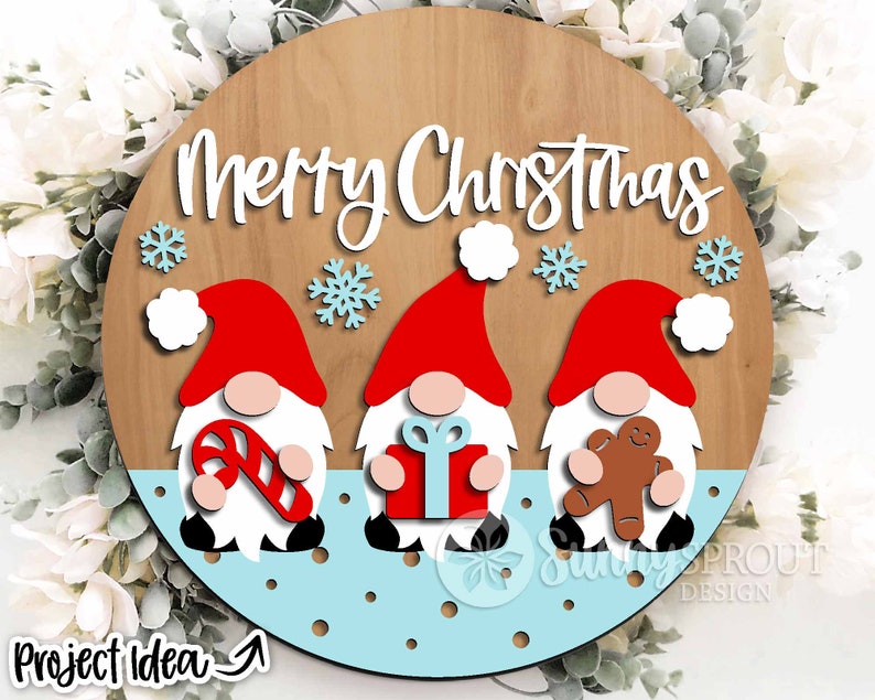 May include: A round wooden sign with the text "Merry Christmas" in white lettering. Three gnomes are depicted on the sign, each holding a different Christmas-themed item: a candy cane, a gift box, and a gingerbread man. The gnomes are red and white with black boots and white beards. The background of the sign is a light blue color with white polka dots.