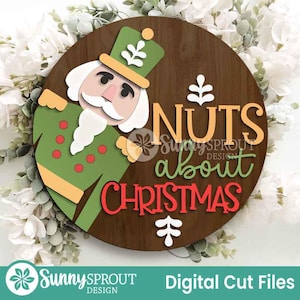 May include: A circular wooden sign featuring a cartoon nutcracker and the phrase "Nuts about Christmas." The nutcracker is green, white, and tan. The text is in orange, green, and red. White flowers surround the sign.