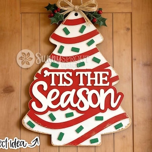 May include: A Christmas tree-shaped wooden door hanger featuring a white background with red diagonal stripes and small green rectangular accents. The text 'TIS THE SEASON' is displayed in white lettering. A jute bow and holly leaves top the piece.