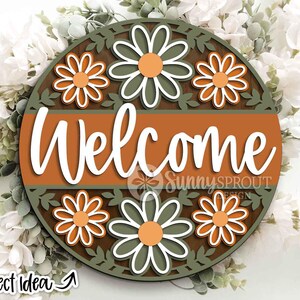 May include: A round wooden sign with the word "Welcome" in white script. The sign features a brown and orange color scheme with daisy flower cutouts and a green leaf border. The sign is surrounded by white flowers and greenery.