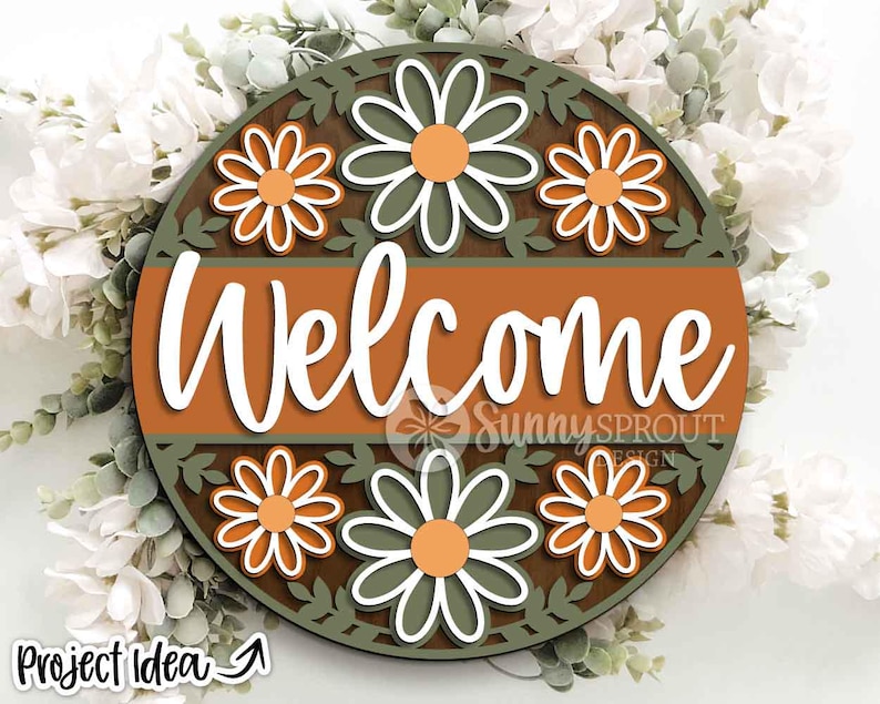 May include: A round wooden sign with the word "Welcome" in white script. The sign features a brown and orange color scheme with daisy flower cutouts and a green leaf border. The sign is surrounded by white flowers and greenery.