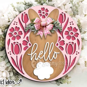 Peut inclure: Panneau de porte de Pâques rond en bois rose avec une silhouette de lapin et des découpes florales. Le mot "hello" est écrit en cursive. Un nœud vichy rose et blanc et de la verdure ornent les oreilles du lapin. Le design comprend des tulipes et des fleurs.