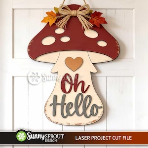 May include: A mushroom-shaped wooden wall hanging featuring a dark red cap with cream-colored spots, a cream stem, and a brown heart. The text "Oh Hello" is displayed in dark red and grey script. A jute twine bow and two autumn leaves sit on top.