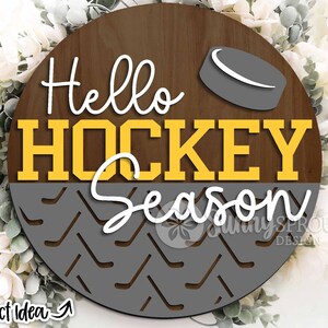 May include: A round wooden sign with the text "Hello Hockey Season" in yellow letters on a brown background. The bottom half of the sign features a gray and white hockey stick pattern.