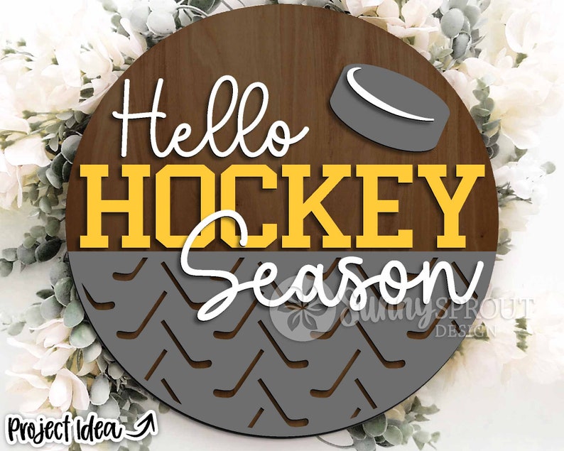 May include: A round wooden sign with the text "Hello Hockey Season" in yellow letters on a brown background. The bottom half of the sign features a gray and white hockey stick pattern.