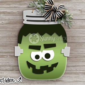 Op de afbeelding: Houten deurhanger in de vorm van een pot met een groen Frankenstein-monstergezicht. Het ontwerp omvat witte ogen, een zwarte mond en een zwart-wit gestreepte strik. De tekst "Sunny Sprout Design" en "Project Idea" zijn ook zichtbaar.